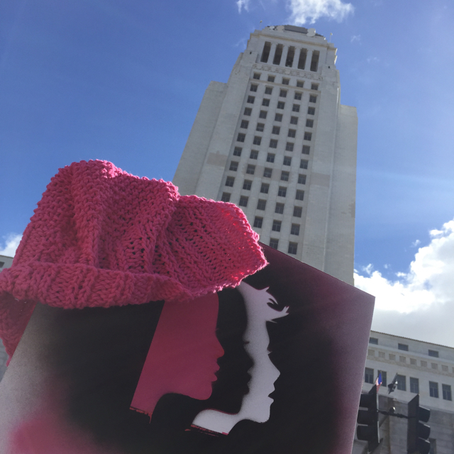 pink knit hat and protest sign with women's march logo held up in front of LA City Hall