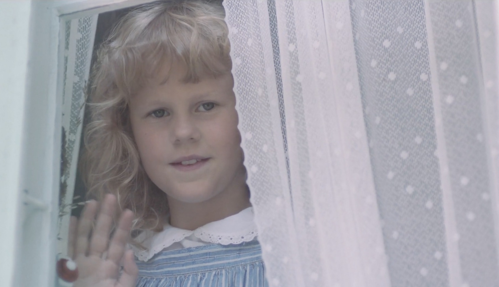 Young girl looks through window with lace curtain and waves