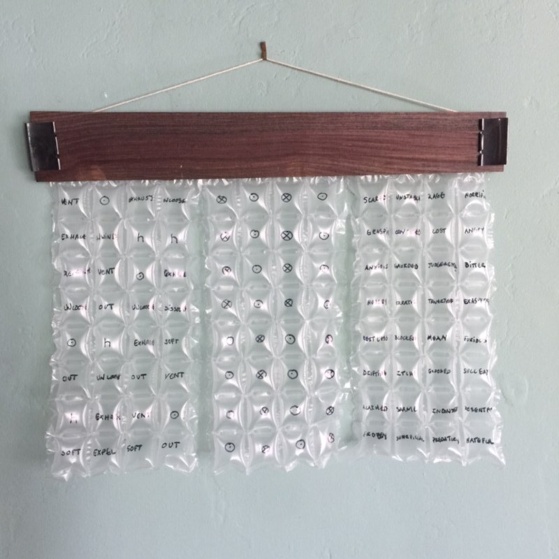 Bubble wrap with words written on each bubble, hanging from a wooden holder secured with binder clips.