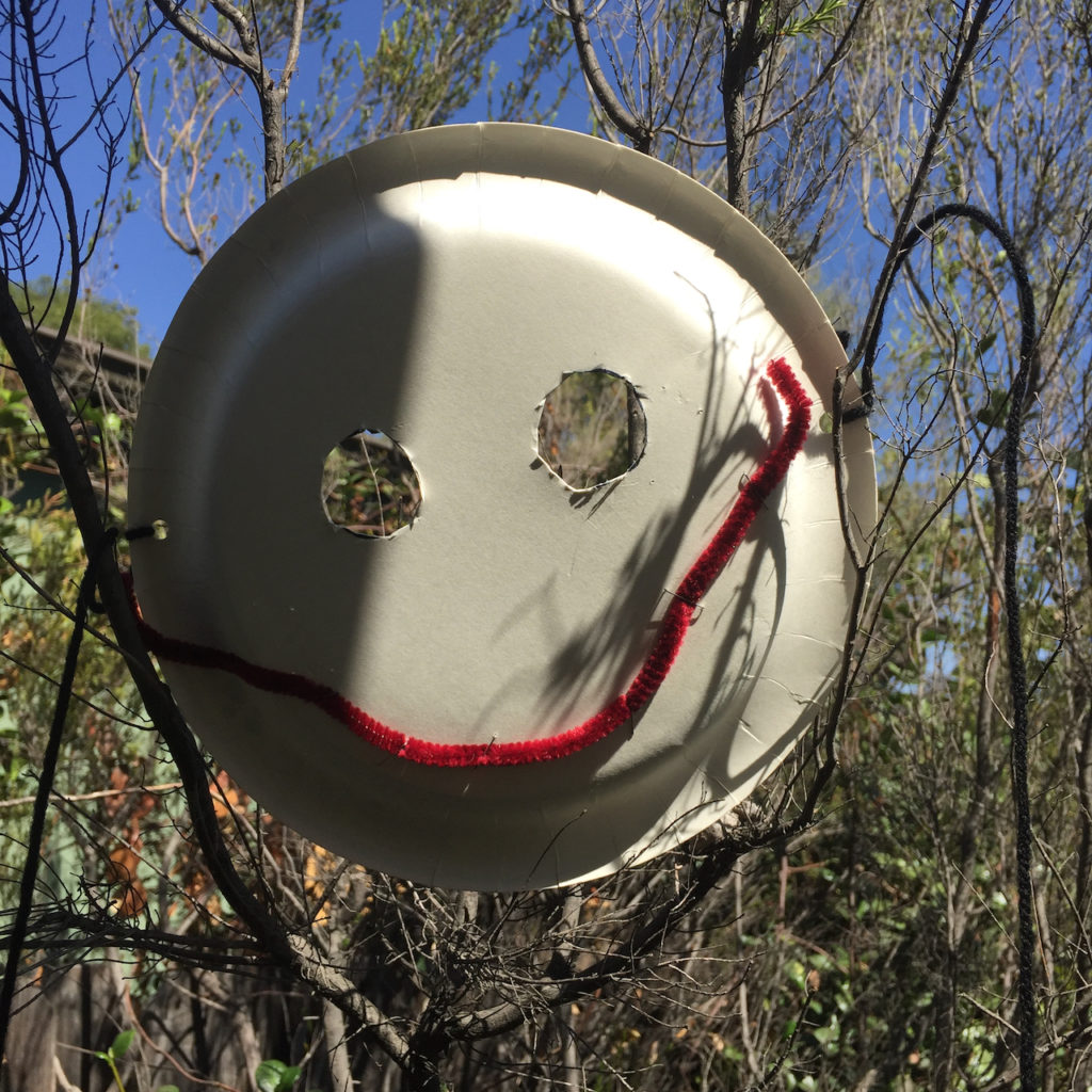 A mask made from a paper plate hung in a tree. The eyes are sloppily cut out and the mouth is maroon pipe cleaner stapled haphazardly in place. 