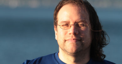 Justin Corwin, head shot, background the blue of the ocean. Sun lighting coming in from the left of the image lighting up the right side of his face.