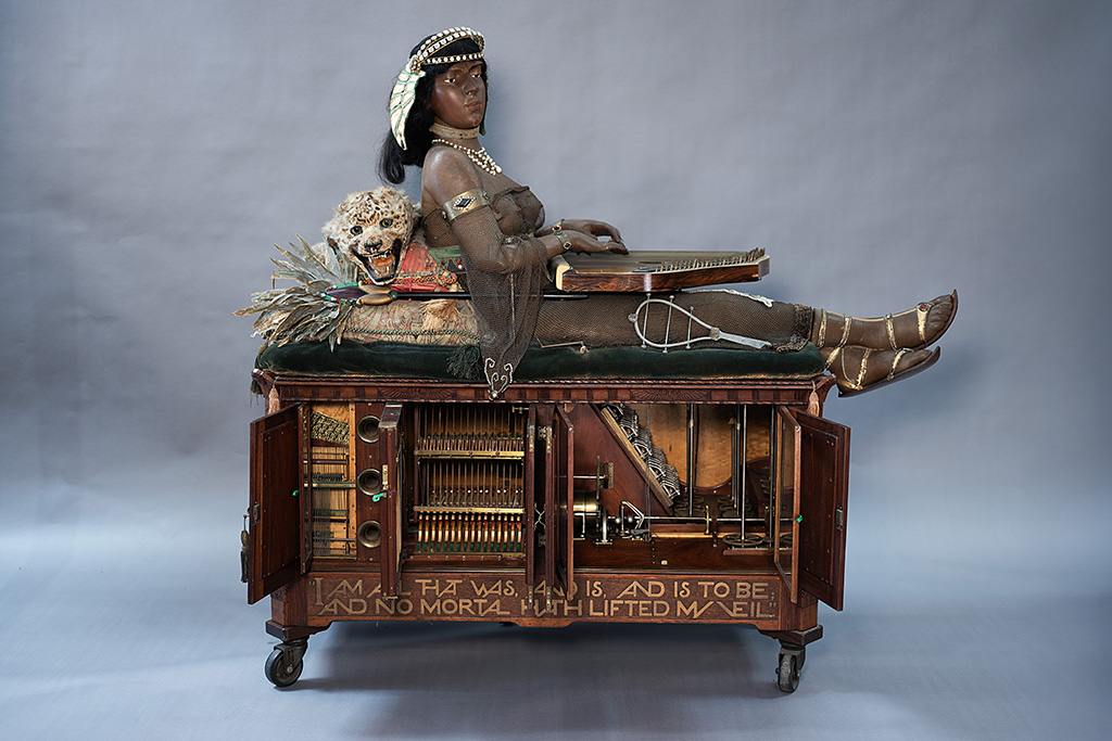 "Isis," a zither-playing automaton. A large wooden box with a variety of open doors, exposing internal mechanisms. On top sits a life-size woman with an ornate headdress, wearing a sheer top and holding a zither. Behind the woman is a mounted large cat