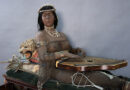 A medium shot of the life-size Isis figure sitting oher cabinet with her zither on her lap and a mounted cat behind her