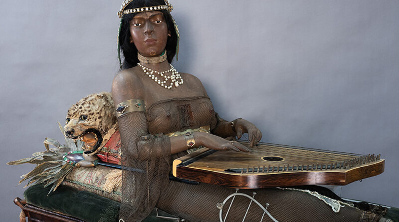 A medium shot of the life-size Isis figure sitting oher cabinet with her zither on her lap and a mounted cat behind her