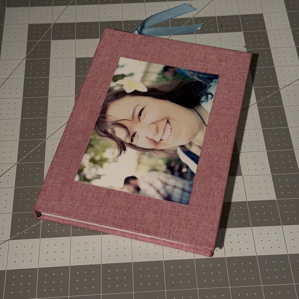 Hardcover book sitting on a gray cutting mat. The book is covered in pink fabric, and has a color photo of a smiling woman with long brown hair, a white/yellow plumeria pinned in her hair. A blue ribbon attached to a bookmark stick out from the top of the book
