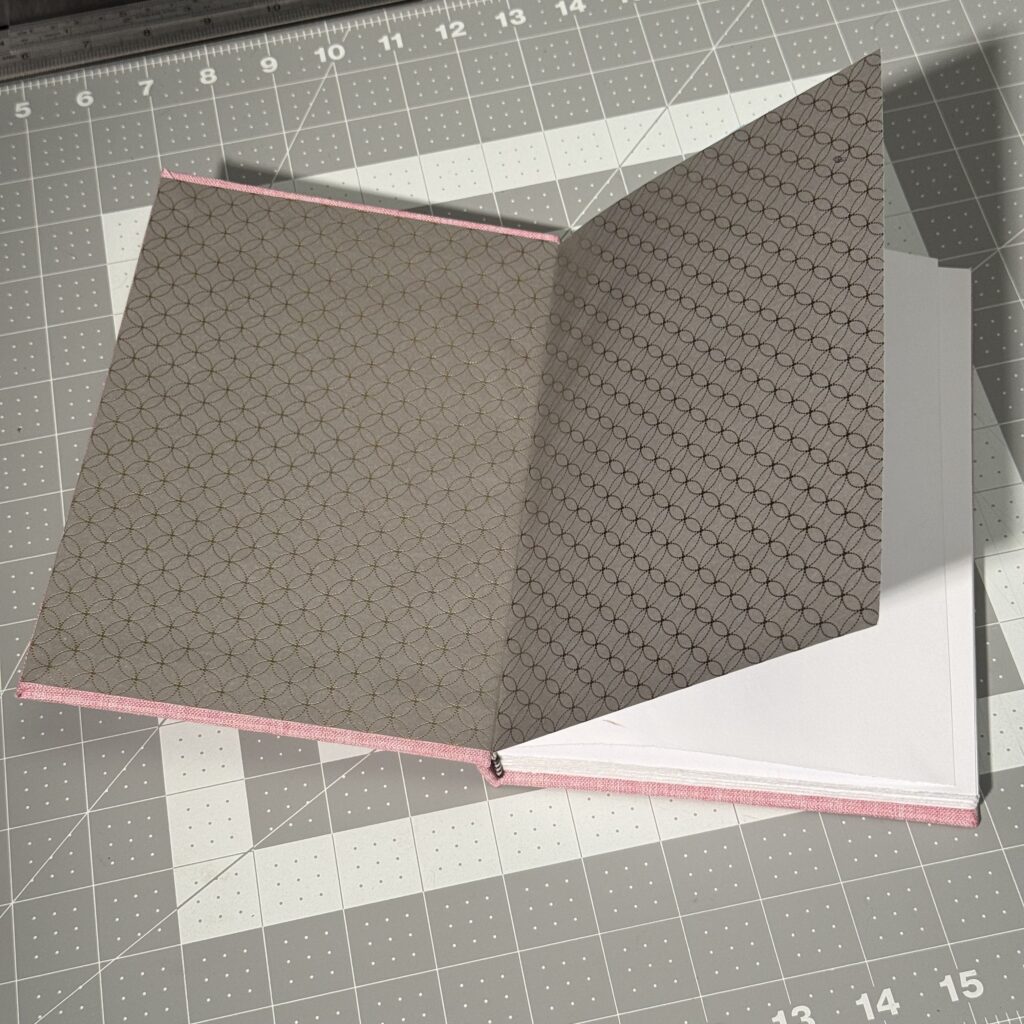 Pink hardcover book on a gray cutting mat, front cover open to show the gray and gold patterned end paper