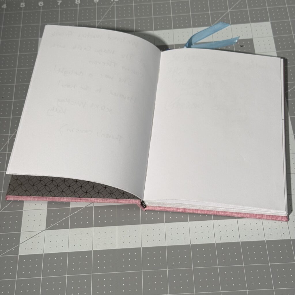 Pink hardcover book on a gray cutting mat, open to show two blank pages. A blue ribbon attached to a bookmark peeks out from the top of the book