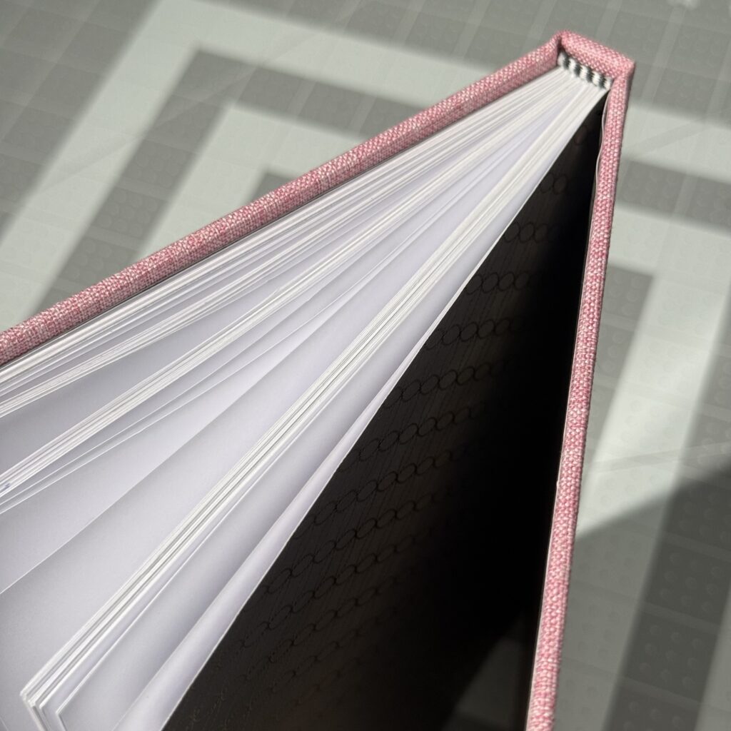The top of a pink hardcover book standing up on a gray cutting mat. The covers are open, and the pages are slightly spread. Black and white headband is visible between the pages and the spine of the cover.