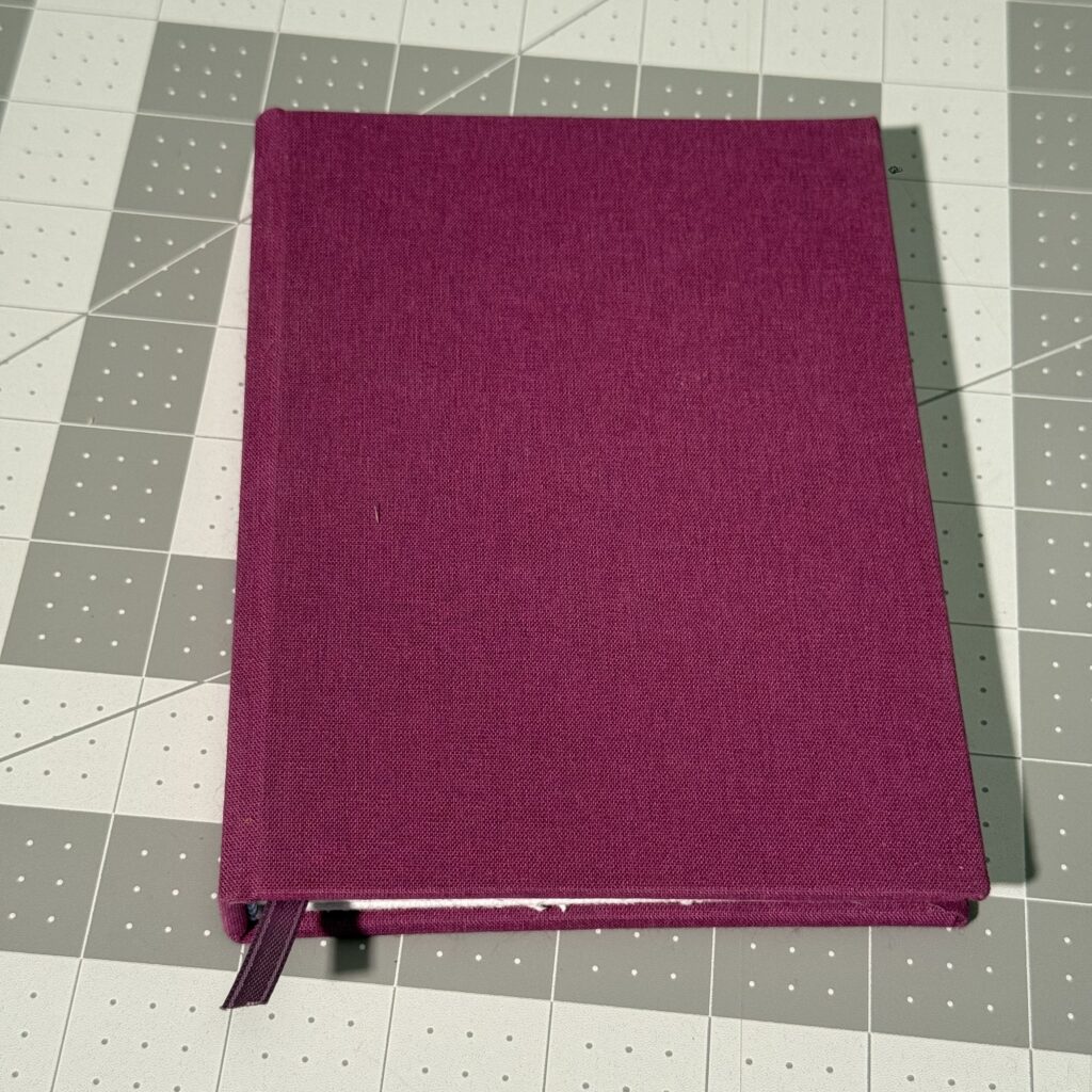 A closed hardcover book sitting on a gray cutting mat. The is covered in purple fabric, and has a matching purple ribbon protruding from between the pages.
