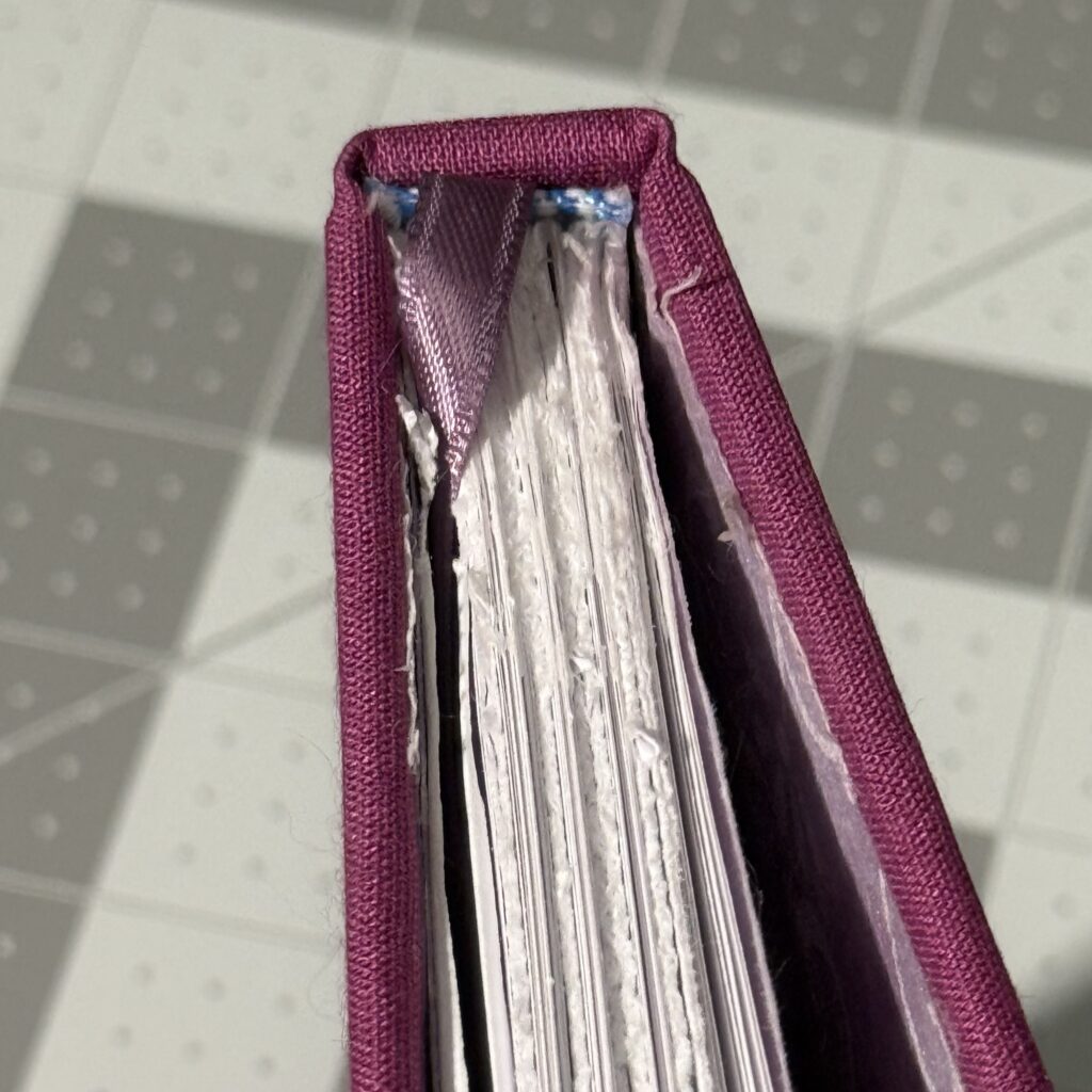 Th top of a hardcover book viewed from above. The book is covered in purple cloth and is standing on a gray cutting mat, and the covers are slightly open, and. the pages are slightly spread. The purple bookmark is seen visible going from the spine to between some of the pages. A blue and white headband is visible peeking out the cover. The pages are roughly cut, and the end paper is roughly attached to the cover.