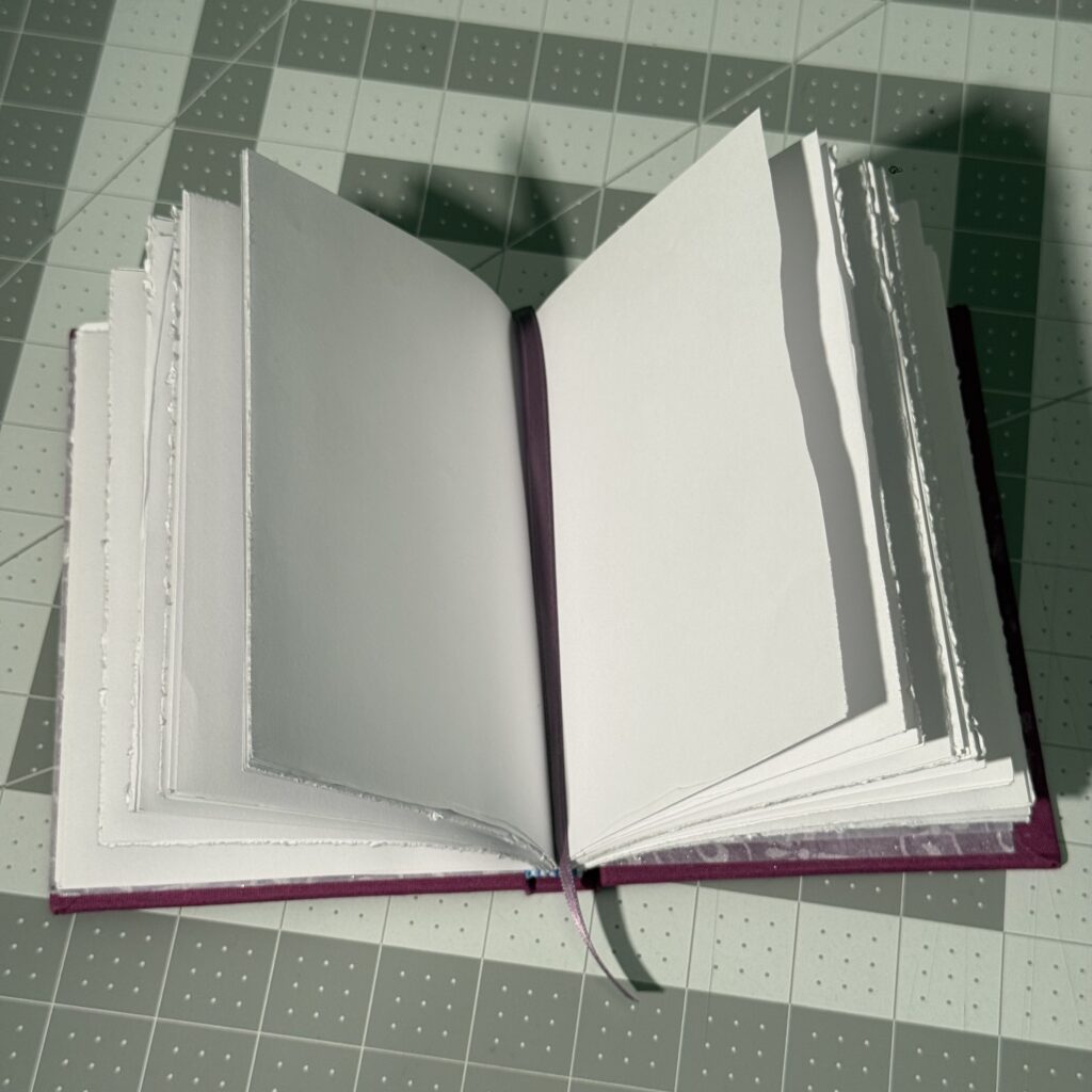 Open book sitting on a gray cutting mat. The pages are spread to two blank pages, and a purple ribbon runs down the page.