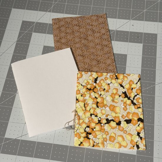 Three small handmade notebooks sitting on a gray cutting mat. Each notebook has different cover - one with a plain off-white cover, one with a gold-toned pattern, and one with a yellow/orange tree pattern.