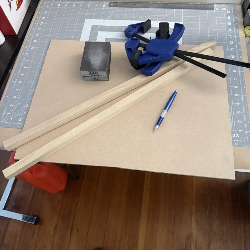 Supplies and tools needed for making the bench hook: a sheet of MDF, two pieces of square stock, clamps, sandpaper blocks, and a mechanical pencil.