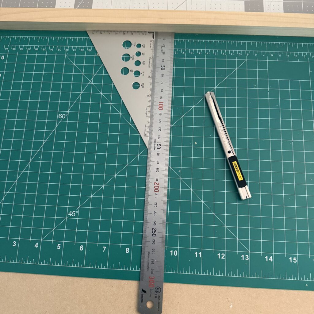 A finished bench hook with a green cutting mat in place, with a metal ruler and triangle placed against the back stop. Next to the ruler sits a craft knife.